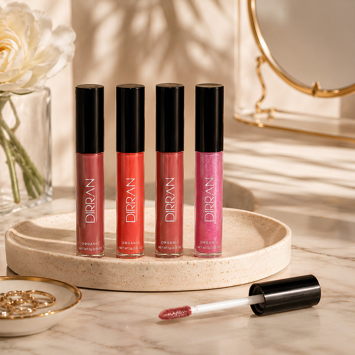 Four DIRRAN Organic Lip Glosses in shades of pink, coral, and berry are displayed on a round beige tray, with one applicator wand lying in front on a marble surface. The scene is styled with a white rose in a glass vase and a gold-framed mirror in the background.