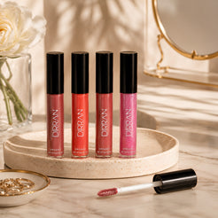 Four DIRRAN Organic Lip Glosses in shades of pink, coral, and berry are displayed on a round beige tray, with one applicator wand lying in front on a marble surface. The scene is styled with a white rose in a glass vase and a gold-framed mirror in the background.