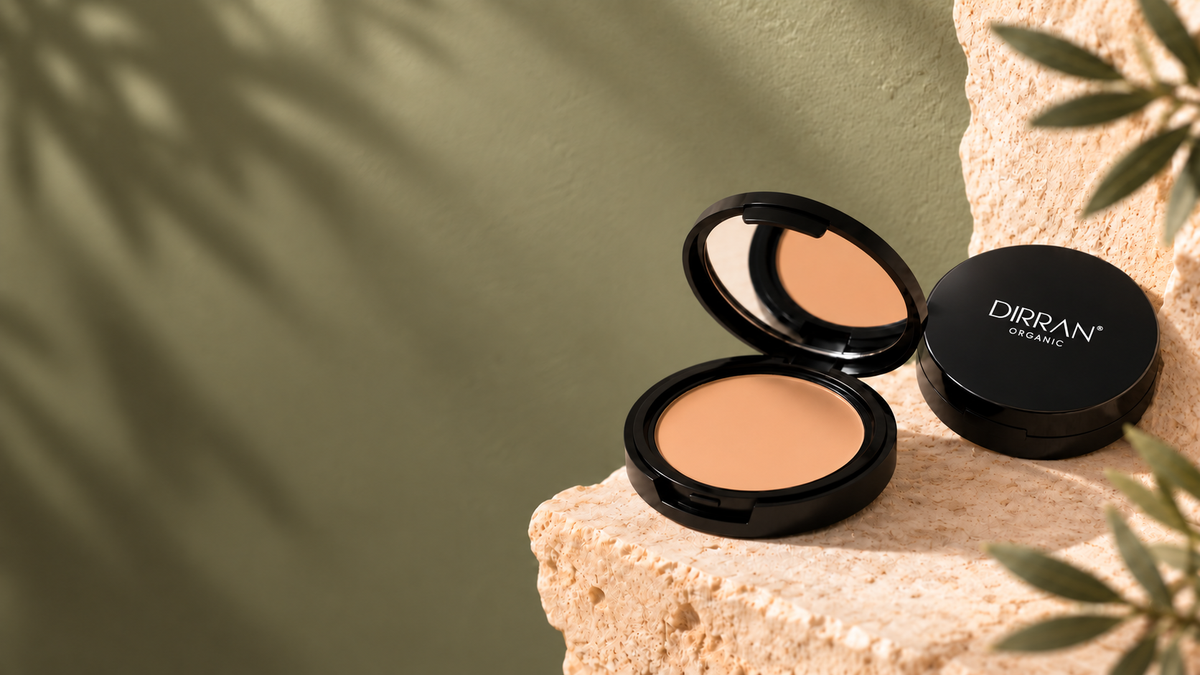 Two black compact makeup cases displaying a warm beige pressed powder, one open and one closed with the brand name DIRRAN COSMÉTICS visible, styled on a textured stone surface against a muted olive green background with soft leaf shadows.